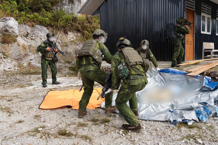 Soldiers in uniform with guns put a dead body on a tarpaulin.