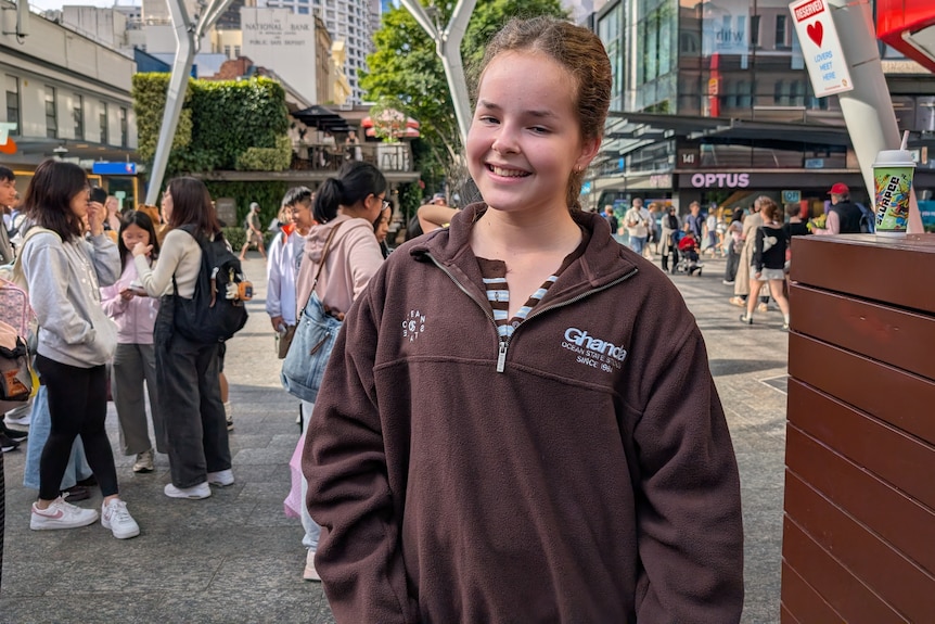 A young girl in a brown jumper smiles widely at the camera with people milling about behind her