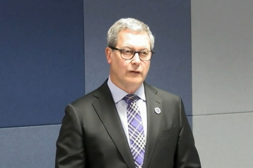 Mr Lancaster in a black suit and purple tie with lines crisscrossing on it standing and asking questions.