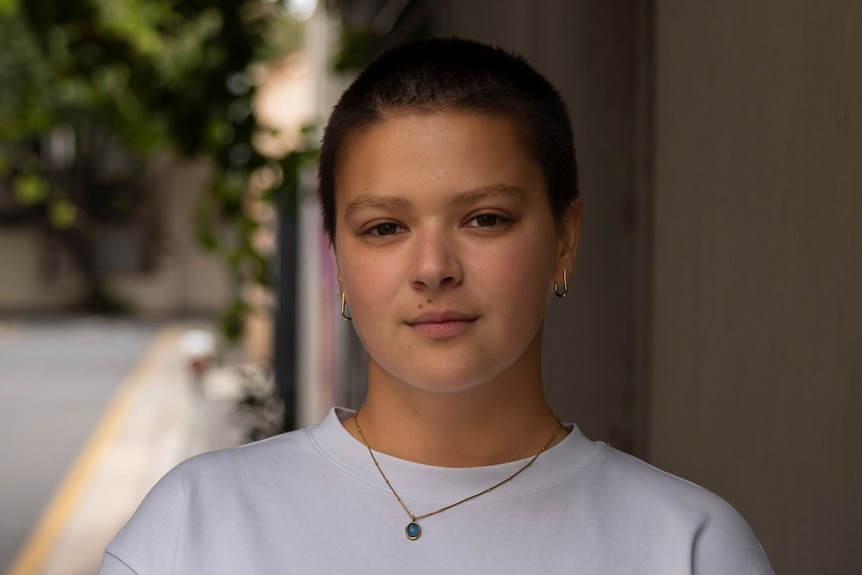 A nonbinary person with a shaved head, wearing a necklace and a white t-shirt.