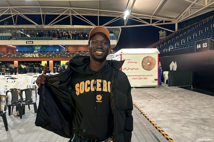A young man smiles. He wears a Socceroos jumper and a black jacket.