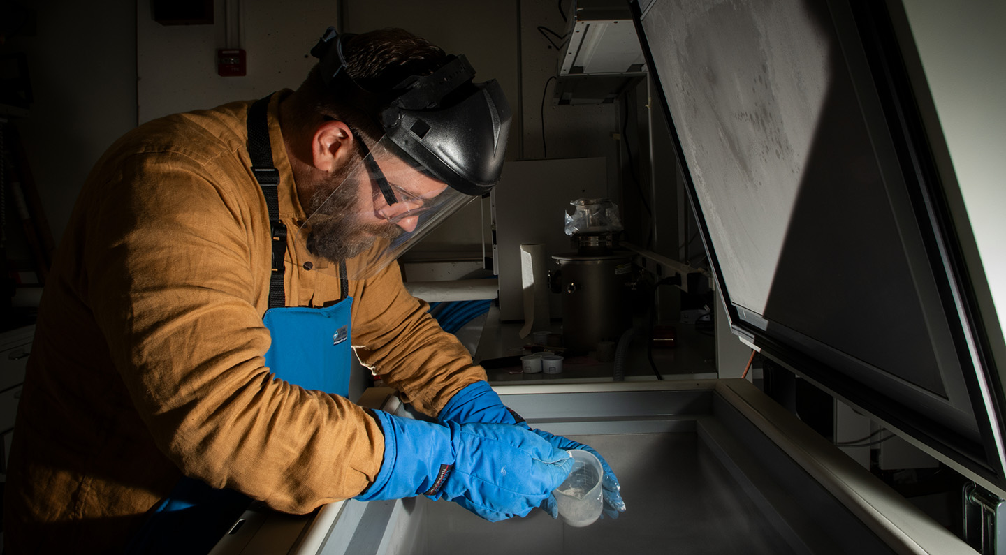 The same man in a light brown shirt, blue gloves, an apron, and face shield works over an open chest container while carefully scooping powdered rock into a small vessel.