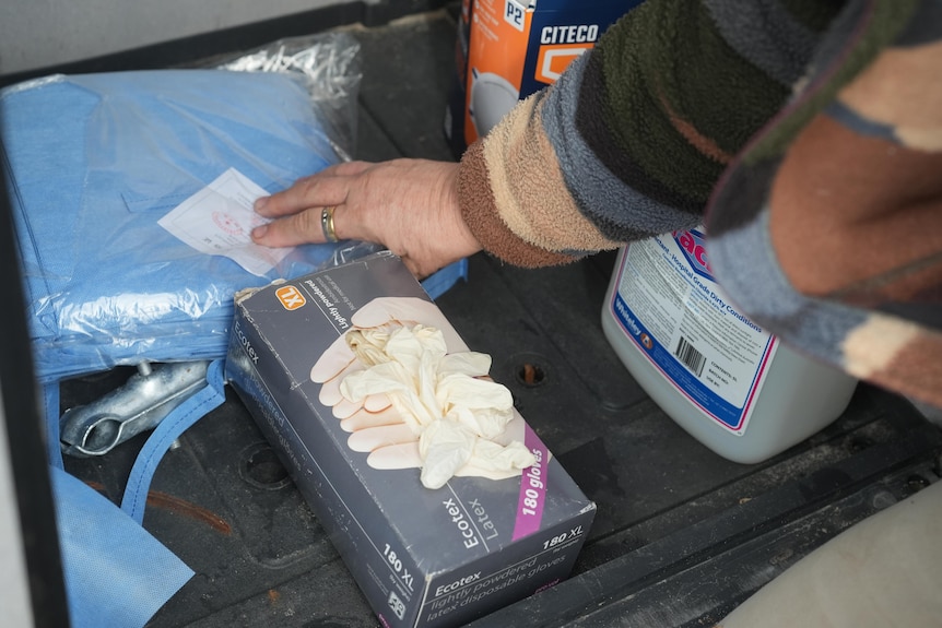 A box of gloves, a packet of blue gowns and a box of masks being reached for by a hand.