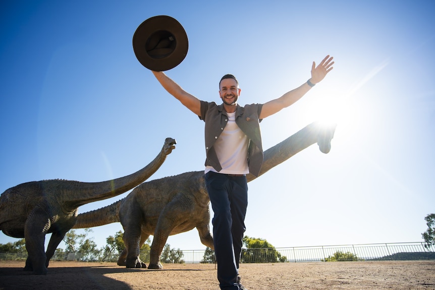 A smiling, dark-haired man holds his hat in the air as he stands near some dinosaur statues in a desert area.