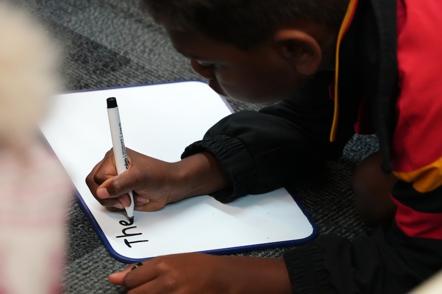 A boy in school uniform sitting on the ground leans over a white board with a marker in his hand.