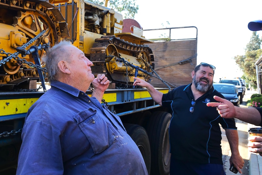 two men gesture towards tractors on the back of a truck