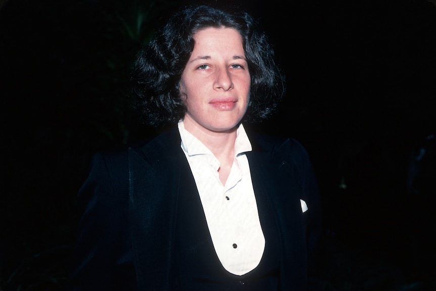 A young Fran Lebowitz gazes towards the camera wearing a black and white three piece tuxedo at night