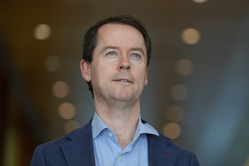 A man with brown hair wearing a blue button up shirt and navy jacket looks up and ahead.