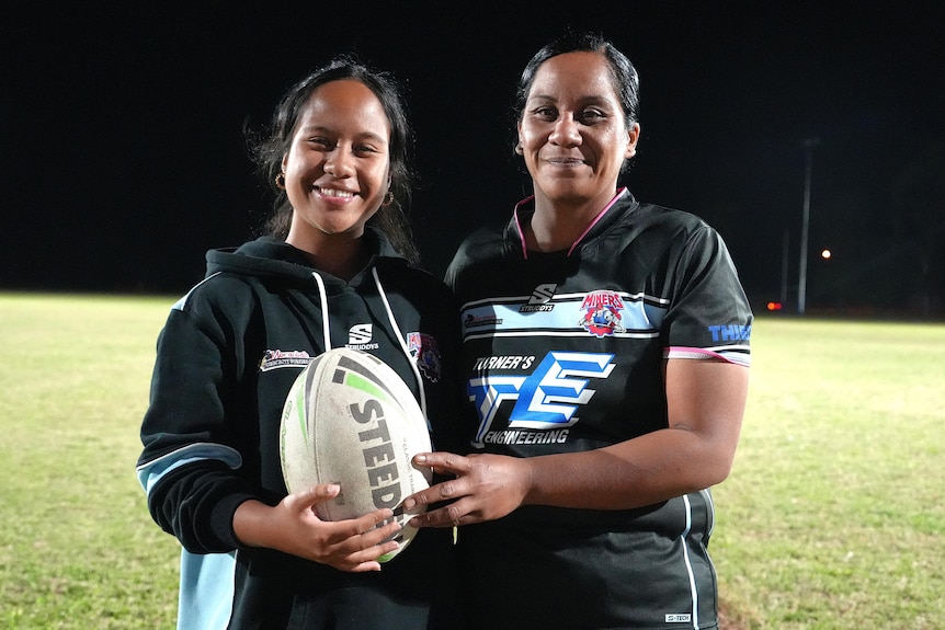 Mother and daughter with football smiling at camera, in uniform, on footy field at night time