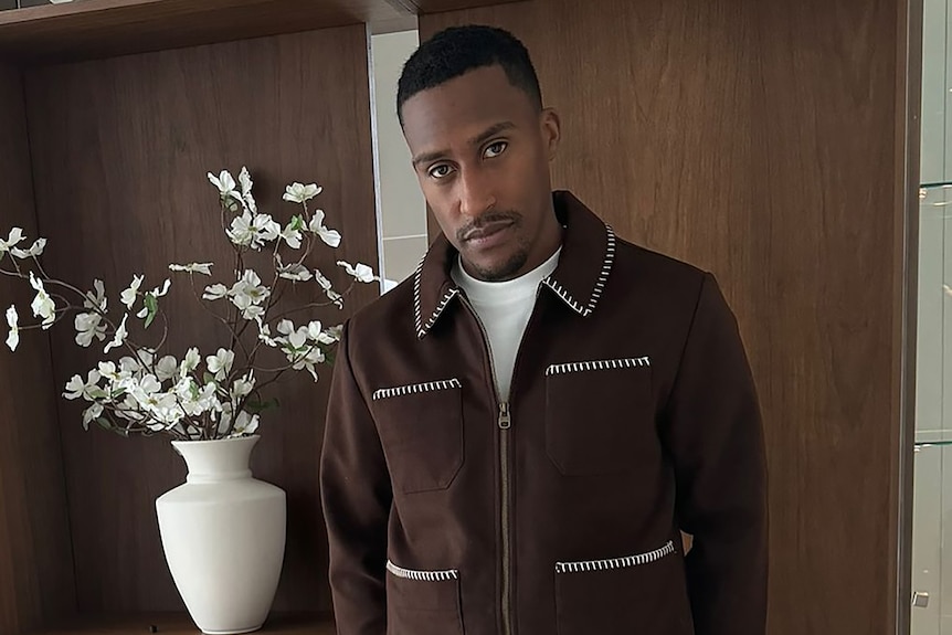 British YouTuber and rapper Yung Filly posing for a photo in a brown jacket, with a white vase and white flowers behind him.