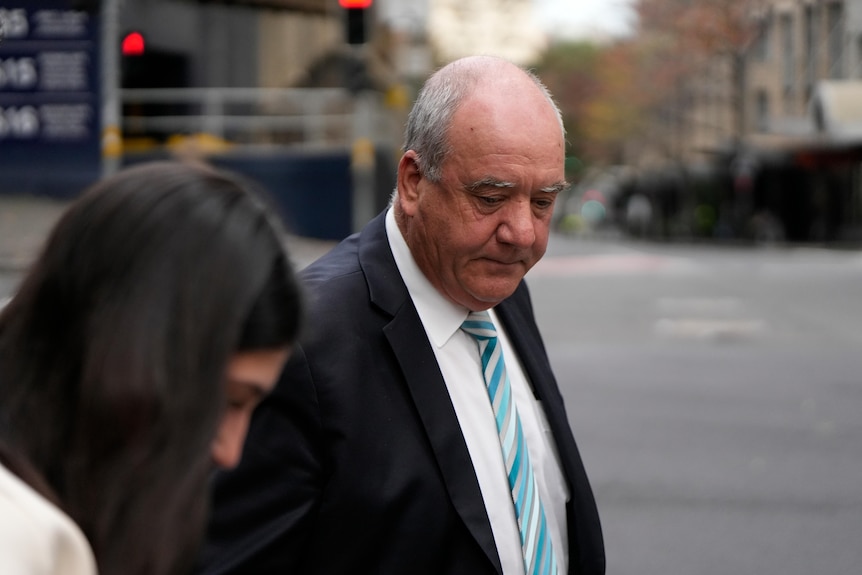 A man looking downwards in a dark jacket, white shirt and tie, crosses an inner city street with a woman with dark hair.