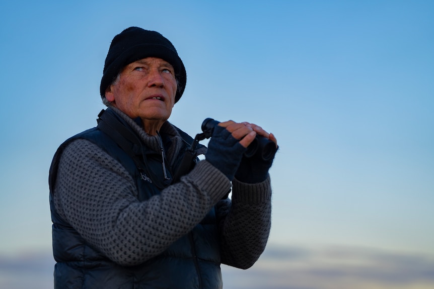 A man with a beanie on stands raises binoculars and looks left of the camera with a concerned look on his face