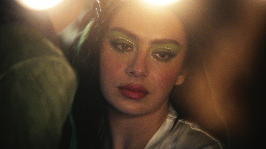 A close up photo of Charli xcx with green eyeshadow and a red lipstick with bright lights above her