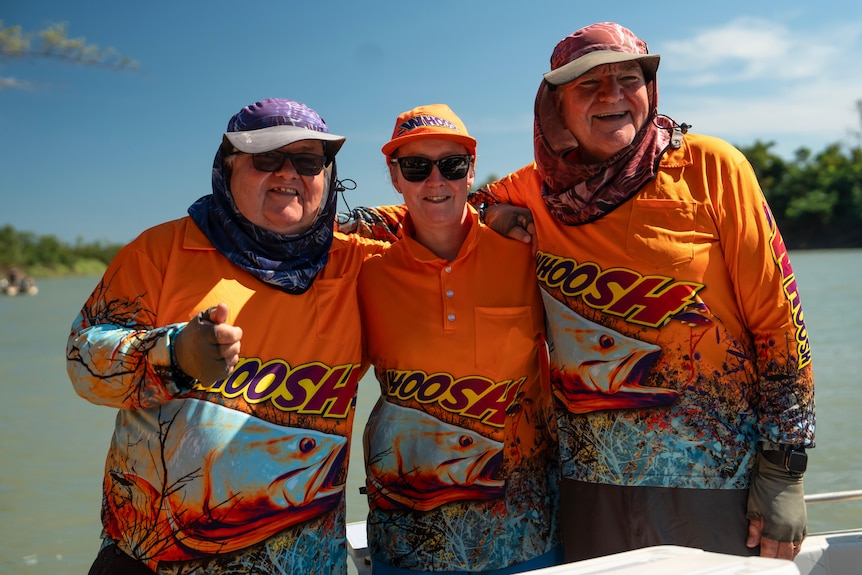 Three people in orange longsleeve shirts standing on a boat, with a river scene behind them.