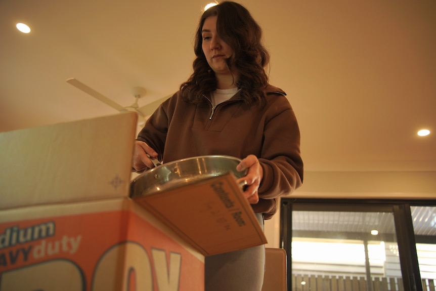 A young lady with shoulder-length brown hair lowers a sauté pan into a moving box in her living room.