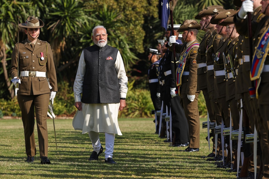 A man in a grey tunic over a white gown walks past a line of soldiers at attention in khaki uniform.