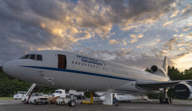 A large white airplane has a white rocket attached to the bottom of its fuselage. There is a small white popup tent covering a section of the rocket, and a person in a red ball cap and blue electrostatic discharge jacket sits in front of it. The rocket has several logos on the side and an American flag painted near the tail. The airplane has a long blue stripe running down the side, under the windows and above the wing. Blue letters spell out “Northrop Grumman” above the windows to the right of the open forward boarding door. Several support trucks and other vehicles are visible in the background. The sky is partly cloudy. 