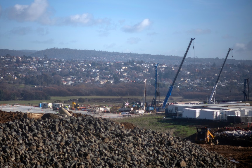Blue cranes at a distant construction site in a valley dotted with homes under a blue hazy sky.