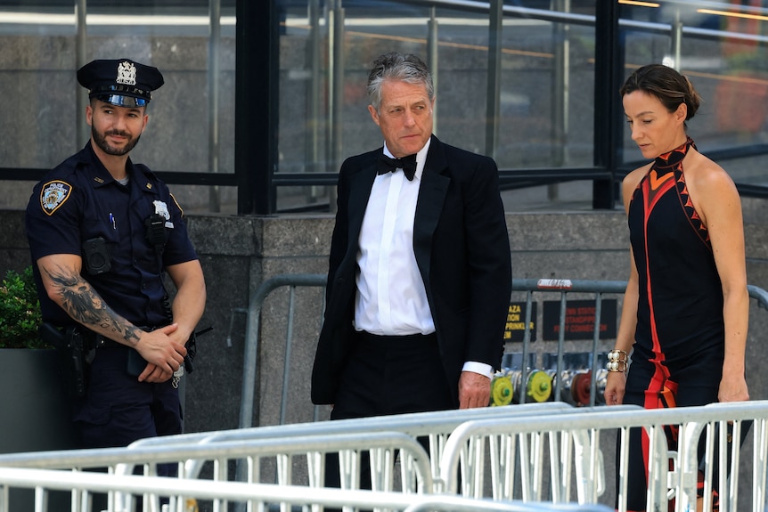 Hugh Grant and partner in a red dress walk down street.