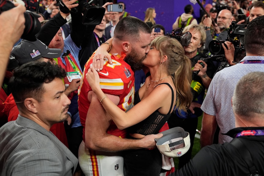 A muscular footballer kisses a blonde pop star as media and camera surround them