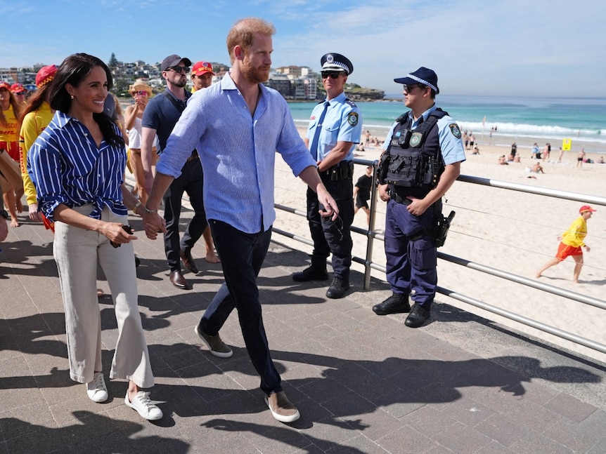 A man and a woman walking near a beach, as multiple people, including police, look on.
