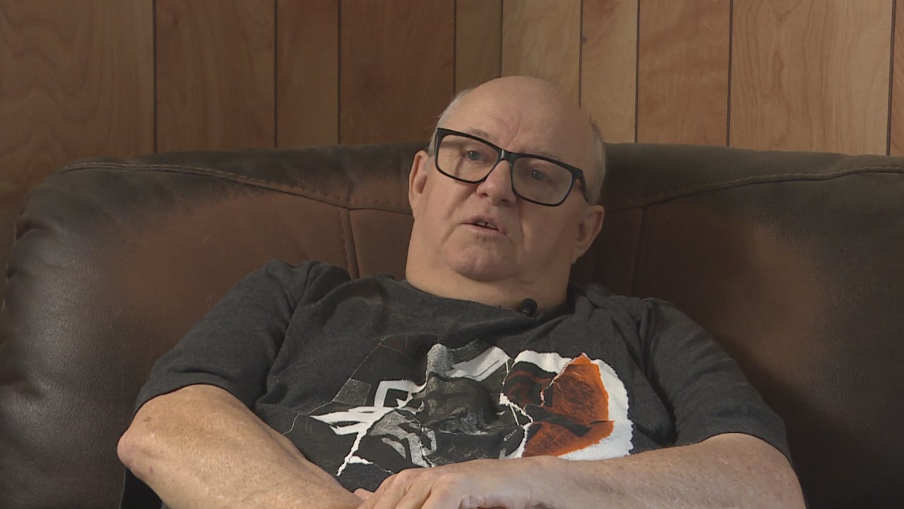 A middle-aged bald man wearing glasses, in a grey t-shirt, sitting on a sofa.