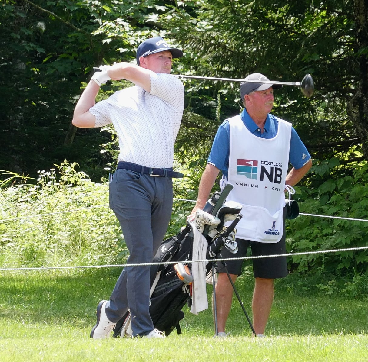 A man swings a golf club on a golf course as an older man stands behind him watching.