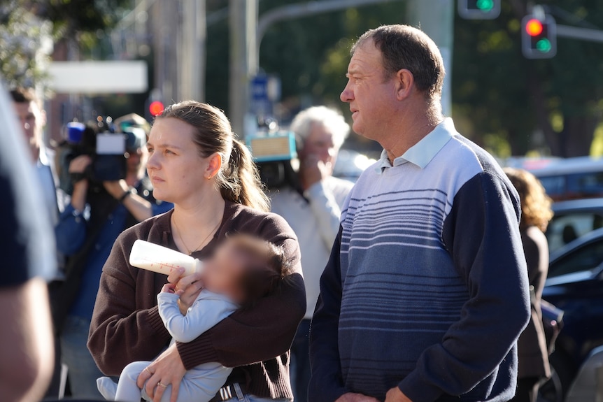 A dark-haired woman feeds a baby with a bottle while standing with a man on a city street.