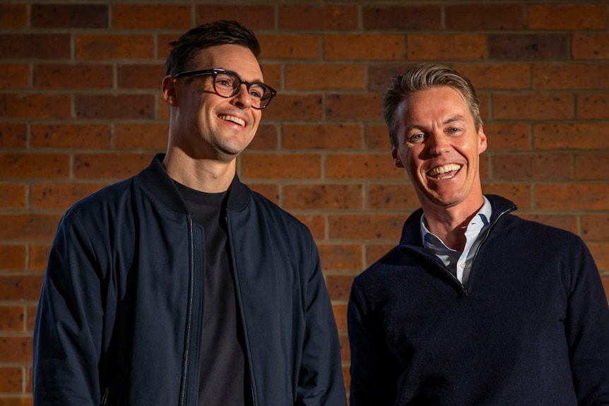two men in dark tops stand against a red brick wall, one brunette and taller, the other with grey hair
