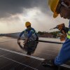 Workers wearing hard hats install solar panels on the roof of a house under a sky filled with gray clouds.