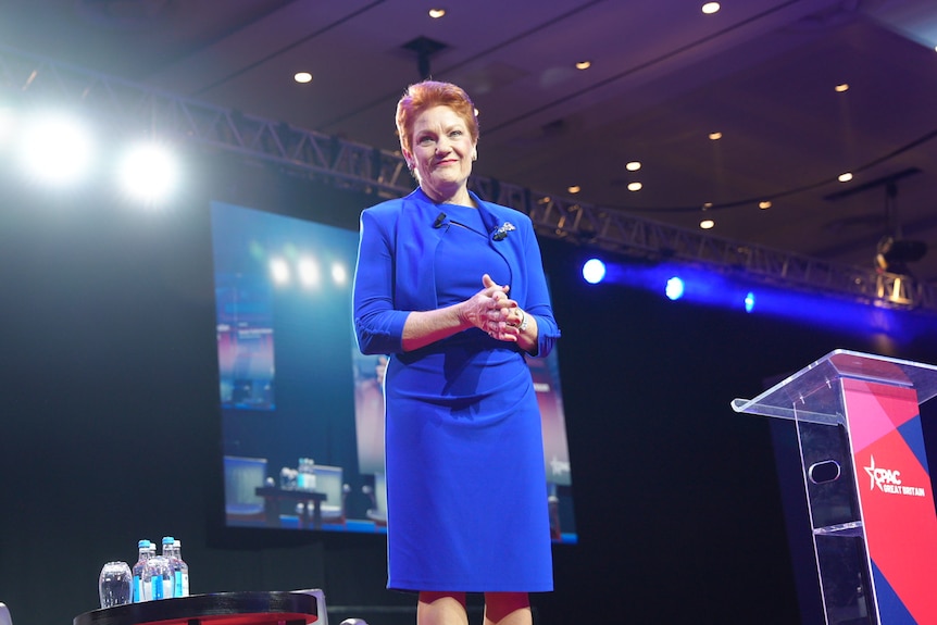 Pauline Hanson wears a blue business dress on stage with a big screen behind her