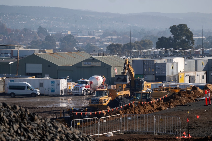 Piles of rocky dirt foregounds a construction site with yellow machinery in a hazy valley dotted with homes.