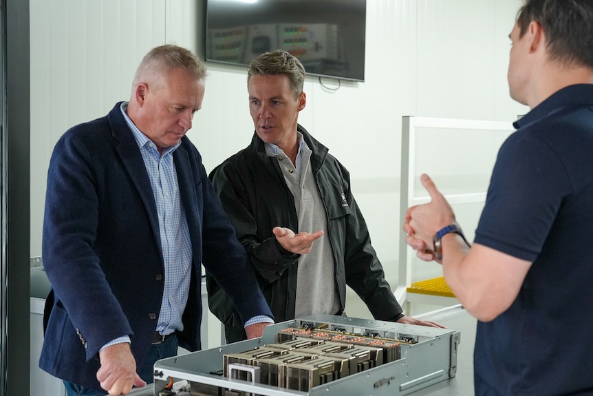 Three men stand over a small model of a building.