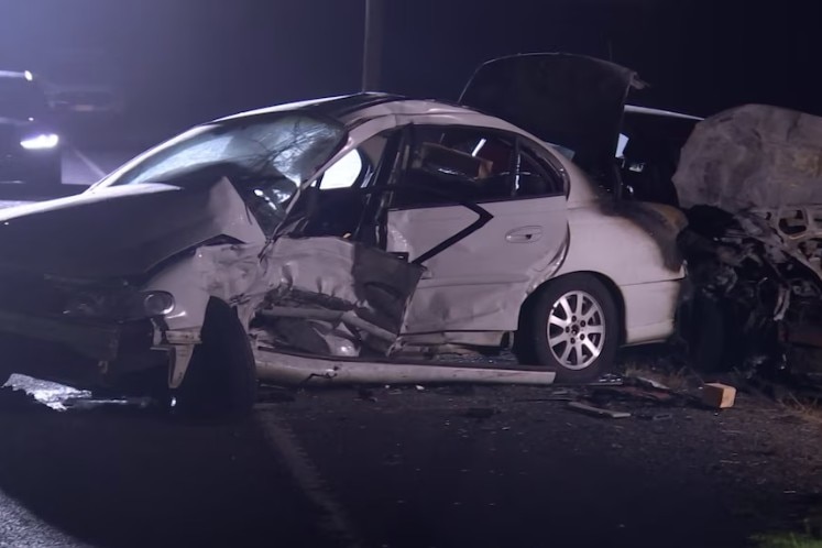 A sedan car is left crumpled on the roadside at night time with an illuminated car in the background.