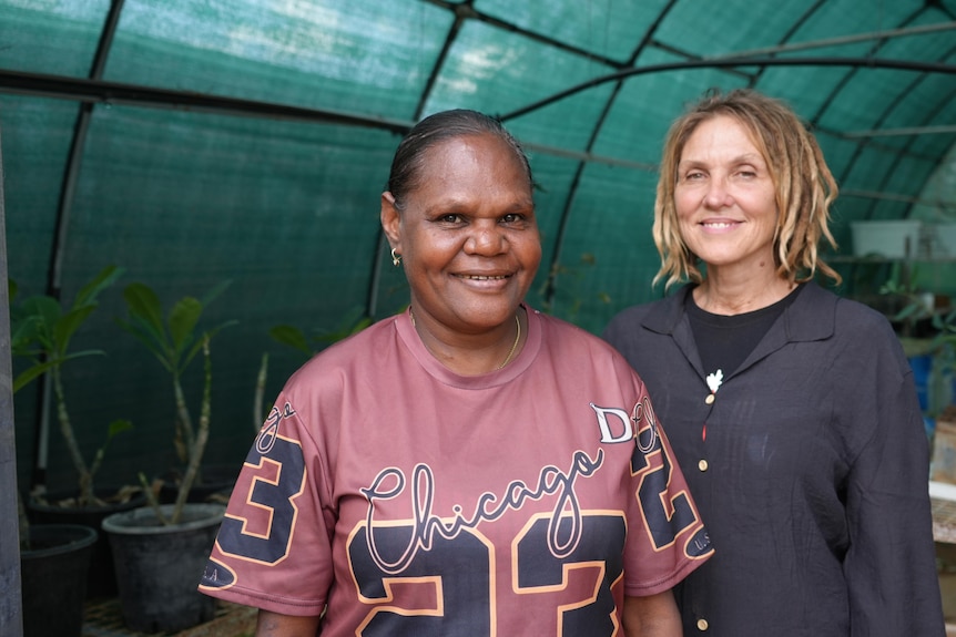 Two women, one Indigenous, one not, stand together in a green house.