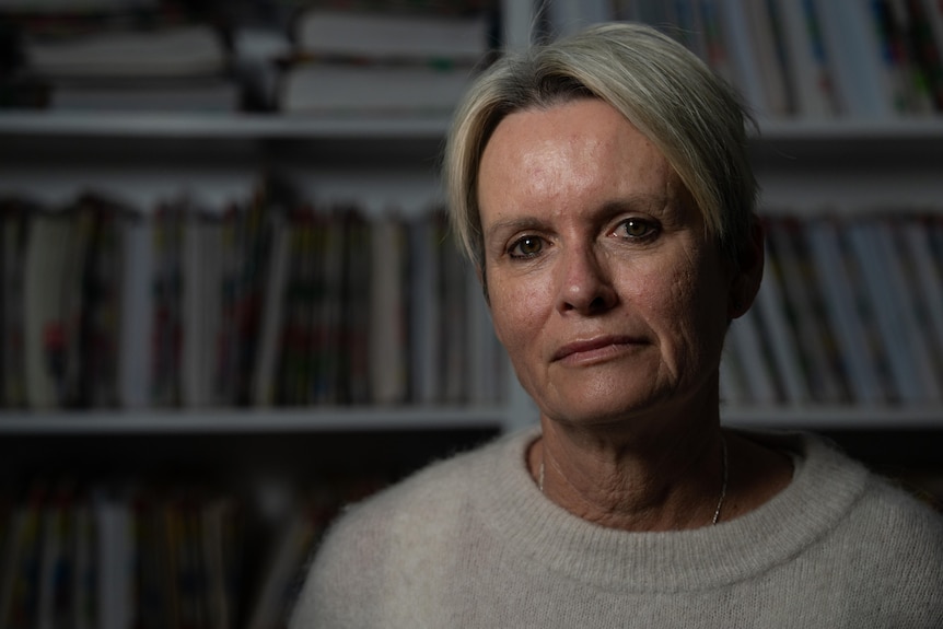 A middle-aged woman with short, blonde hair sits in front of bookshelves in a dimly-lit room.