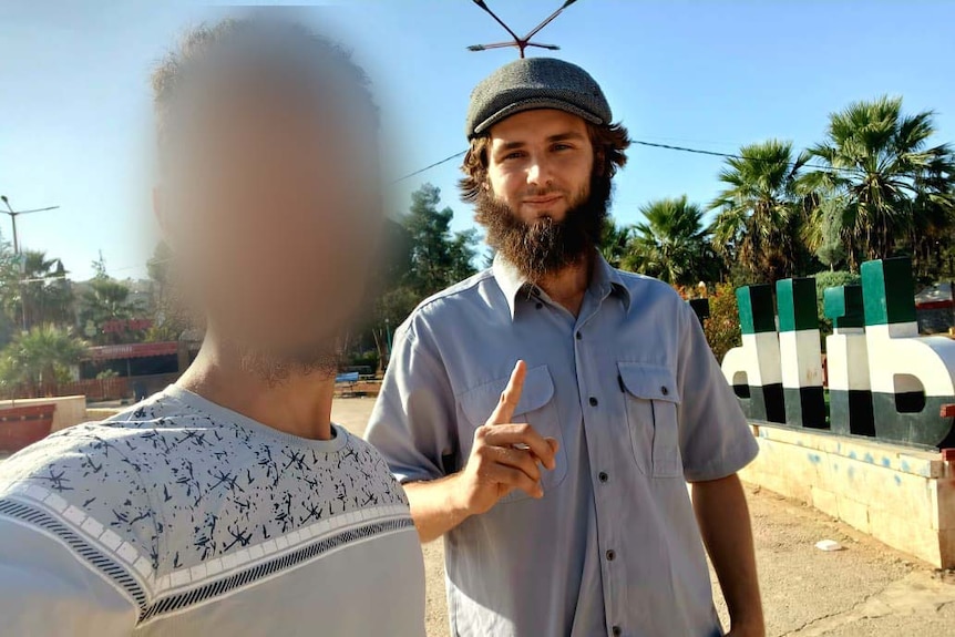 A young man with a beard raises his index finger while posing with another man, who is blurred.