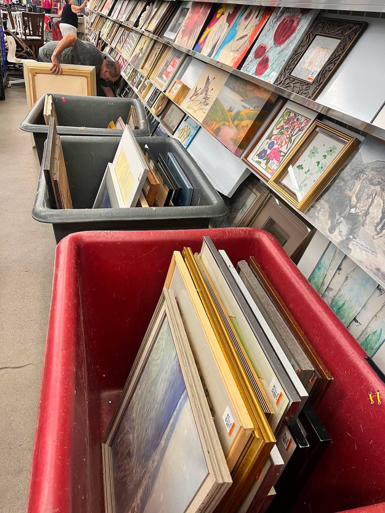 Photo inside a Value Village art aisle. Framed pictures line wall shelves. Large plastic bins are in front of the shelves. They are filled with more framed pictures.