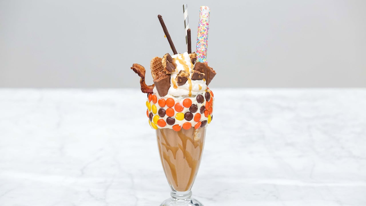 A milkshake in a glass with candy on the rim and chocolate bars sticking out of whipped cream on top.