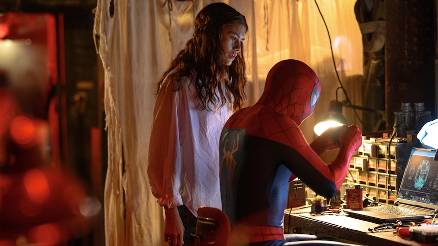 MJ stands over spiderman who is on a computer