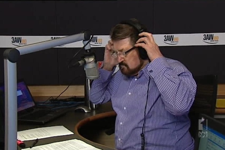 A dark haired man with a beard and sunglasses puts headphones on as he sits behind a microphone.