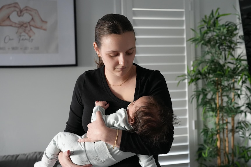 A dark-haired woman cradles a baby in her arms while standing in a living room.