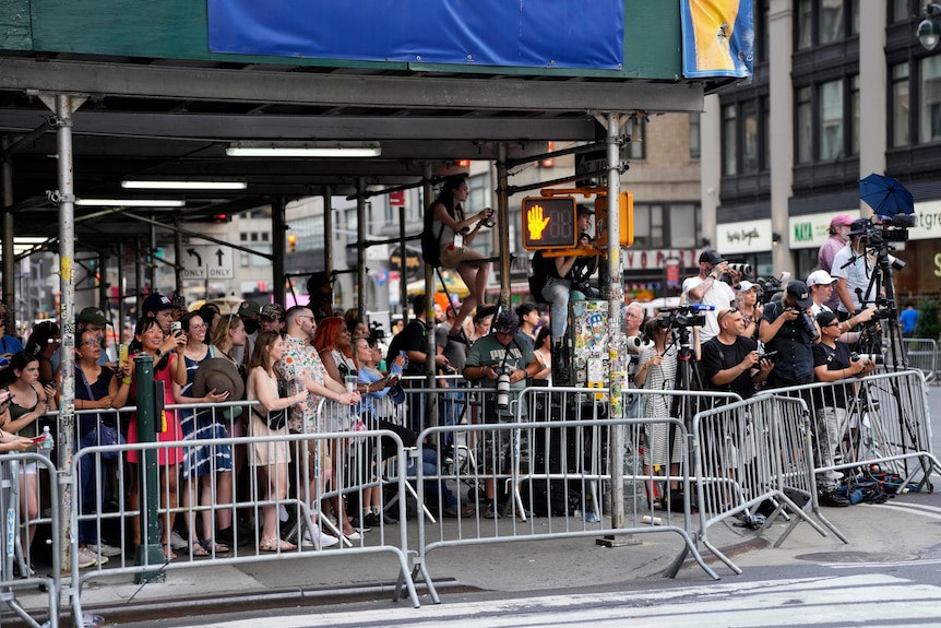 A queue of fans and media stand on a sidewalk.