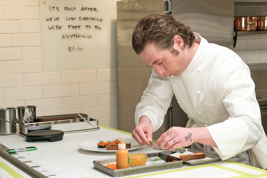 A young white man in a white chef's jacket, stands in a busy commercial kitchen finessing a dish.