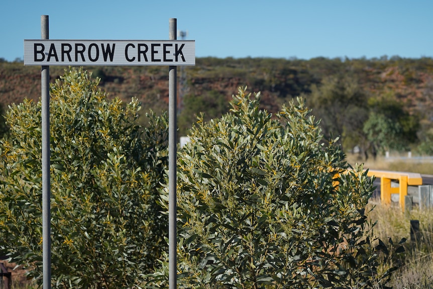 An outback landscape runs along a highway, a roadside sign in the tall grass reads: BARROW CREEK