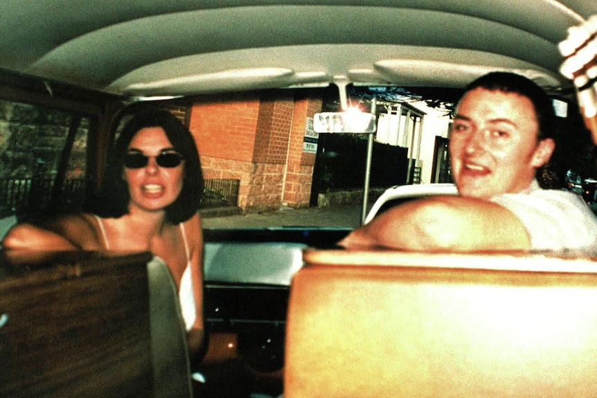A old photo of couple sitting in the front seats of a Kombi van, they are turned around to look at the camera.