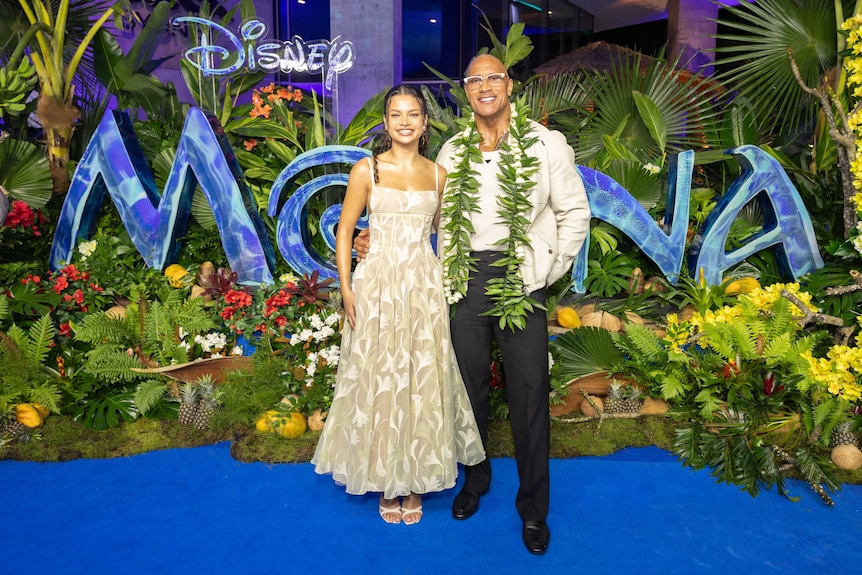 Catherine Lagaʻaia and Dwayne Johnson standing in front of a Moana sign with flowers and plants surrounding them