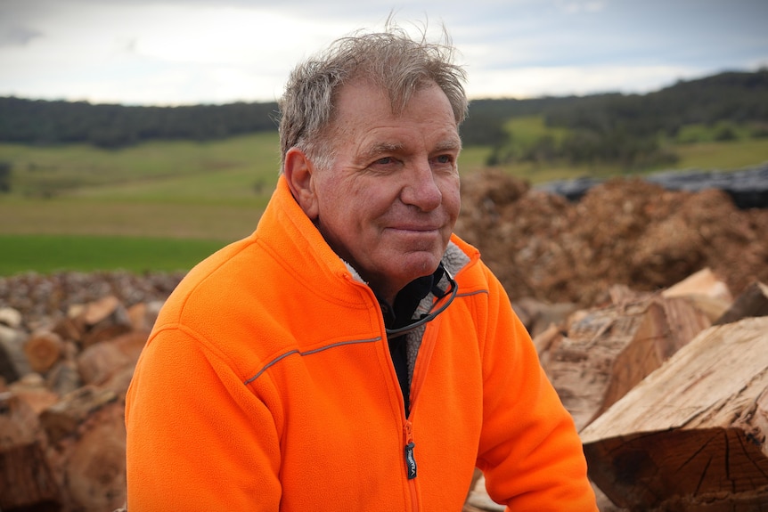 Man in orange high-vis jacket, grey hair, sitting on pile of firewood, farm in background
