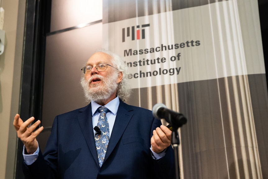 A bearded man with white hair and glasses in a blue suit, mid-speech in front of a microphone, before a sign for MIT.