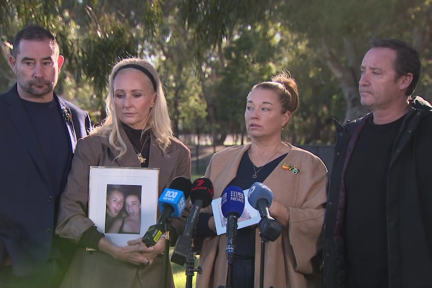 Two  men in dark jackets and shirts booked two women in beige coats and black tops who stand behind microphones.